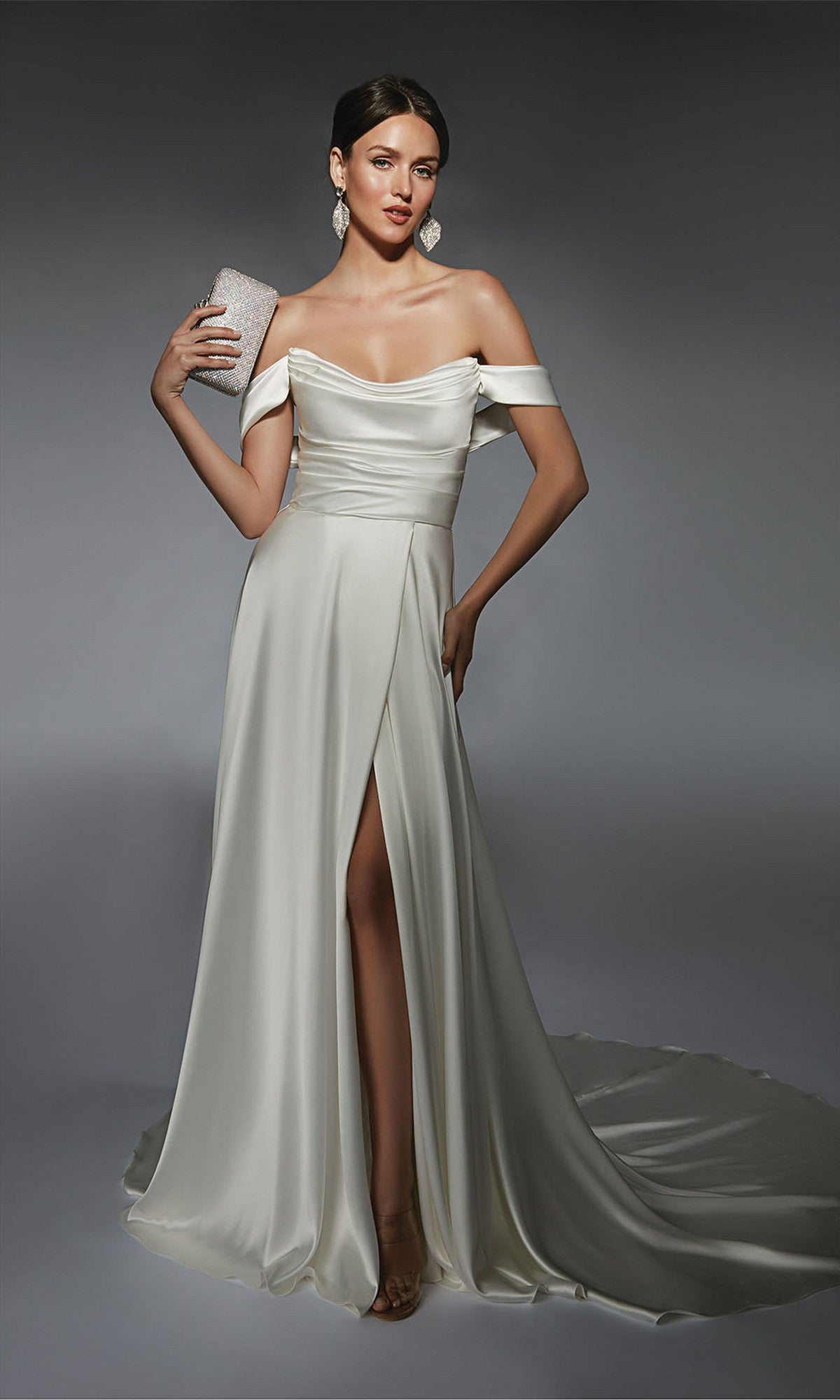 Model in the Alyce 7107 long white dress, featuring an off-the-shoulder neckline and thigh-high slit in diamond white, holds a silver clutch while posing against a plain gray background.